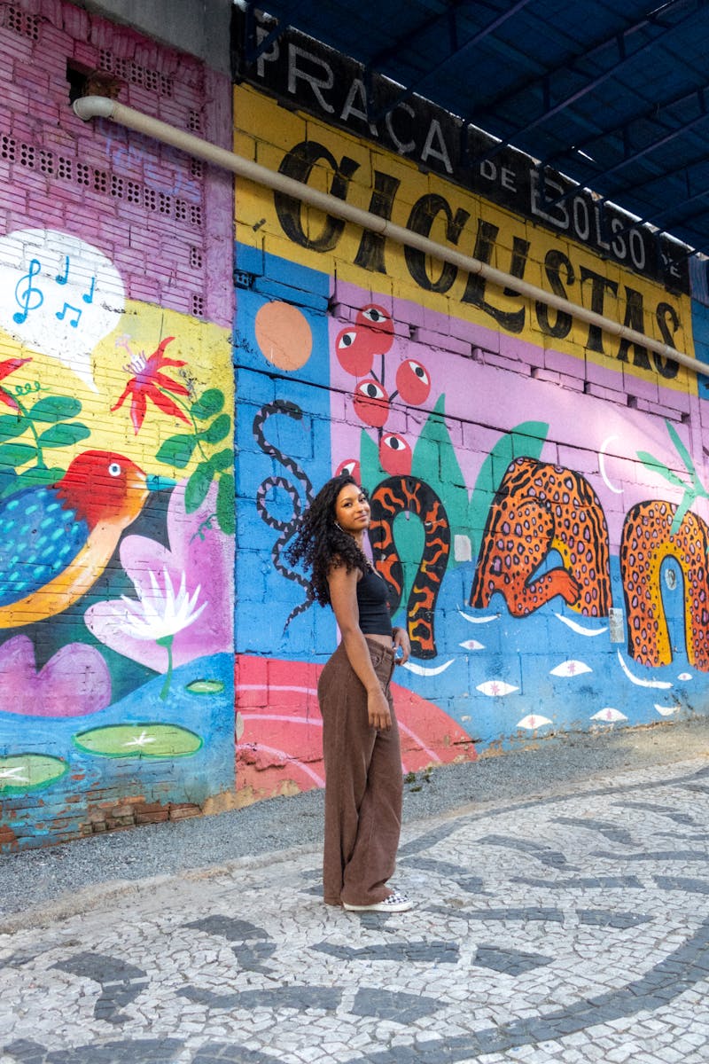 Une femme élégamment vêtue se tient devant une fresque murale vibrante avec des éléments d'art urbain dans une rue brésilienne.